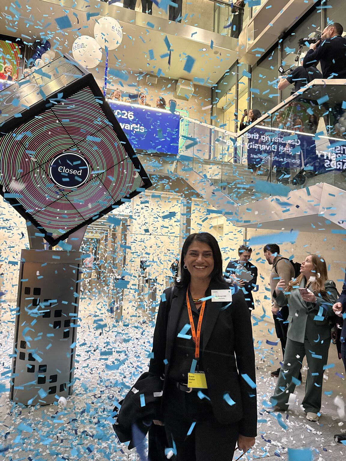 Tab Ahmad OBE, wearing a black blazer and an event lanyard, smiles warmly as blue and white confetti falls all around her. To her left is a large, angled digital display showing the word "Closed" within colorful, concentric rings, resembling a stock exchange closing event. In the background, people can be seen watching and taking photos from the glass balconies of a modern, multi-level atrium.